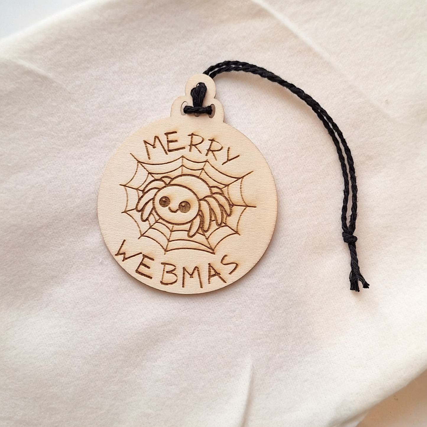 Wooden ornament with 'Merry Webmas' text and spider design on a white fabric background