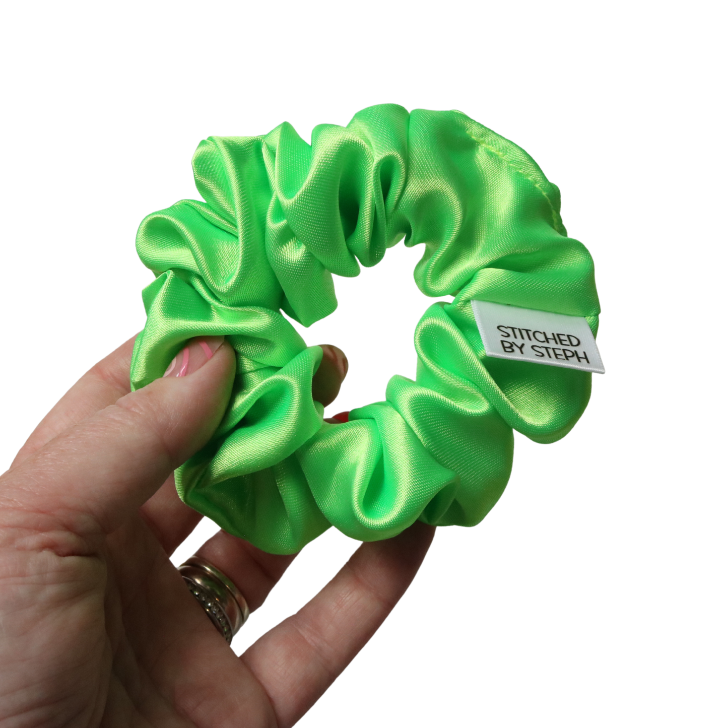 A bright lime green satin hair scrunchie held by a hand against a plain white background. A small white fabric tag on the scrunchie reads 'STITCHED BY STEPH'.