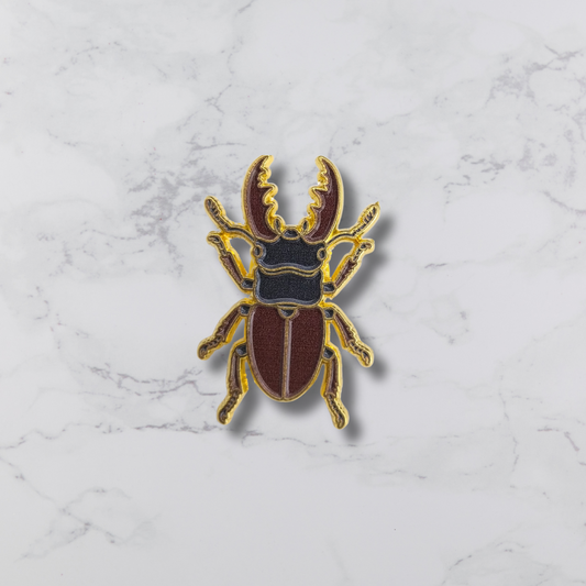 A beetle-shaped enamel pin with gold metal outlines, a dark red body, and a black thorax, centered on a white marble background.