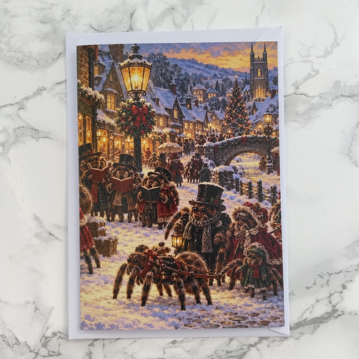 A Christmas-themed art print or card featuring a snowy village scene at night with glowing street lamps, warm window lights, a Christmas tree, and large spiders in the snow, displayed on a light marble surface.